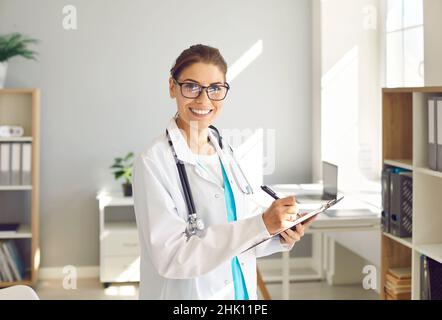 Portrait of happy female doctor in glasses taking notes on clipboard and smiling Stock Photo
