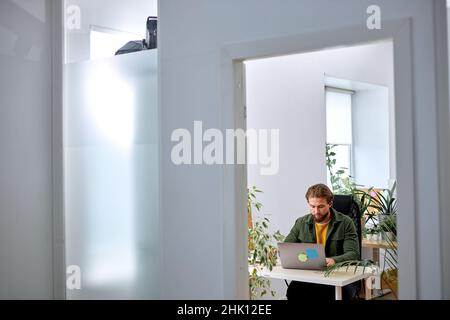 Analyzing data on laptop. young concentrated bearded man in casual wear sit with laptop behind desk in modern office, looking at screen, typing, worki Stock Photo