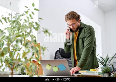 young bearded man standing behind table and talking on phone. Businessman is serious and concentrated. There are laptop and cup of coffee at table. Ha Stock Photo