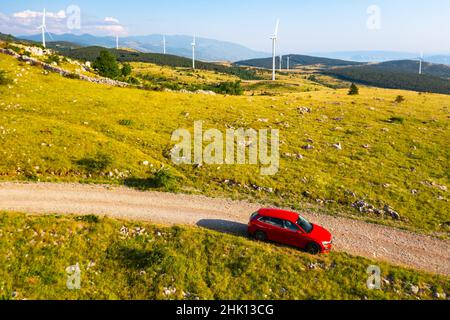 Red car drives on ground road running past windmill farm providing green energy in highland against forestry mountains on sunny summer day aerial view Stock Photo