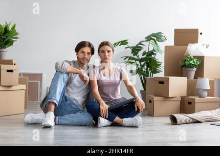 Serious millennial european guy and lady sitting on floor among boxes and plants in room, take a break Stock Photo