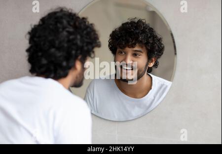 Portrait of happy indian man looking at his reflection in round mirror on the wall, young guy standing in bathroom Stock Photo