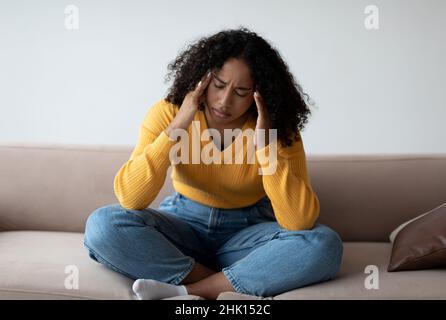 Young black woman with depression touching her temples, feeling stressed on sofa at home Stock Photo