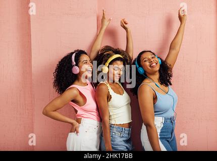Side view of a group of young women raising hands up. Three females wearing headphones while standing against a pink wall. Stock Photo