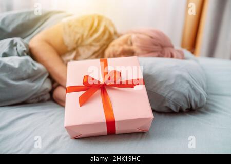 Woman in sleep mask sleeping in bed near paper gift box with red satin ribbon decor. Christmas, New Year, Valentine's Day and birthday concept Stock Photo