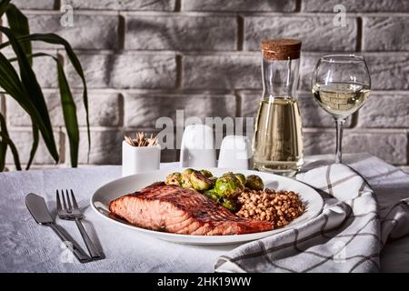grilled salmon fillet with roast brussel sprouts and cooked farro on a plate with restaurant table setting, white wine in glass and in decanter and lu Stock Photo