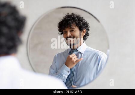 Handsome indian businessman putting on necktie and looking at his reflection in the mirror, standing in bathroom Stock Photo