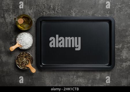 Empty baking sheet, olive oil jar, salt and pepper grains over black textured background. Rectangular oven tray and spices for baking and roasting. Stock Photo