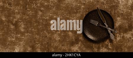 Knife and fork crosswise on a plate. Elegant vintage knife and fork crossed on a black dinner plate over textured background. Sepia toned wide image. Stock Photo
