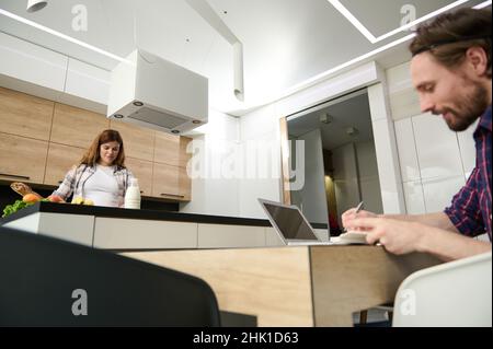 Happy couple at home. Beautiful pregnant woman preparing meal in the kitchen while her husband working on laptop remotely Stock Photo