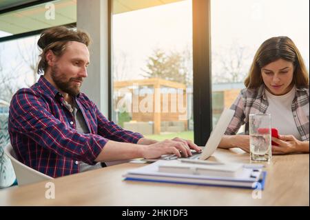 Handsome Caucasian man, entrepreneur, developer, freelancer, working remotely on laptop, sitting at a table next to his wife, who uses a mobile phone Stock Photo