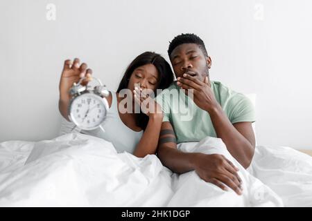 Tired sad sleepy millennial african american couple sit on white bed in bedroom, yawn and show alarm clock Stock Photo