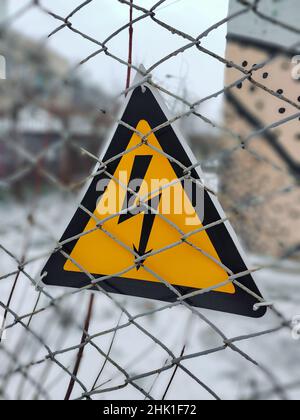 A triangular warning sign hangs on a wire fence about dangerous high electrical voltage behind it. Stock Photo