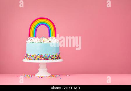 Blue Birthday Cake with meringue decorations, sprinkles and a colorful homemade rainbow topper over a pink background. Stock Photo