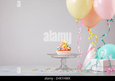 Rainbow Birthday cupcake on a stand with presents, hats and colorful balloons over light grey background. Scene from a birthday party! Stock Photo