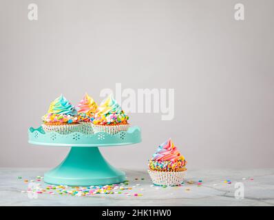 Rainbow Birthday cupcakes with colorful sprinkles on a blue cake stand over light grey background. A delicious dessert. Stock Photo