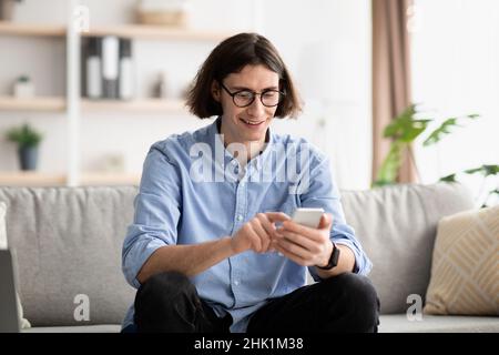 Cool gadget and application. Young man holding cellphone, typing message and surfing internet, sitting on sofa Stock Photo