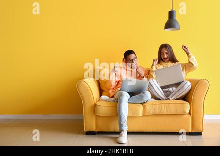 Happy young couple with laptops sitting on sofa near yellow wall Stock Photo