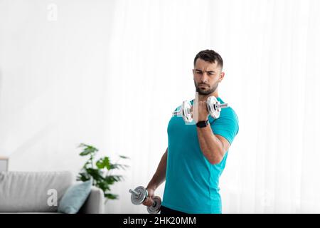 Serious young european guy doing strength exercises for arms, lifting dumbbells in room interior, on window background Stock Photo