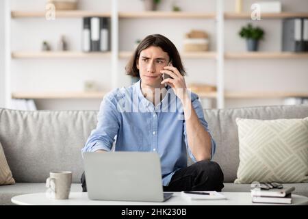 Computer problems. Displeased young guy talking on smartphone and using laptop, calling customer support hotline Stock Photo
