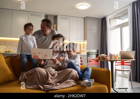 Mother sitting with son on sofa and enjoying daughter drawing Stock Photo