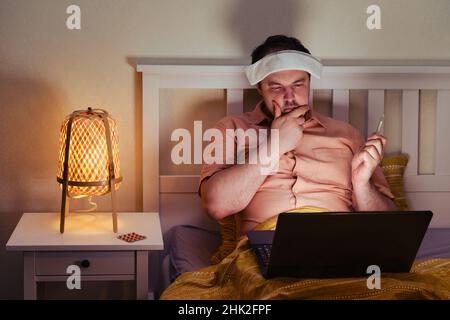 An adult sick man with a pensive face works at a laptop on a bed in the light of a night lamp Stock Photo