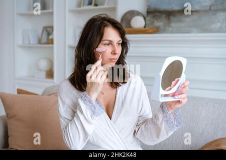 Woman using face roller. Sculpting, lifting and anti-aging care. Skin care and beauty at home. Woman using calming quartz roller massager while sitting on sofa in living room looking in mirror Stock Photo