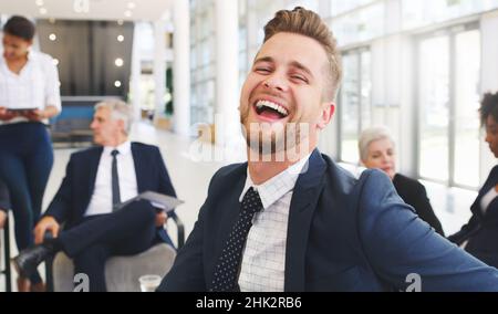 Smiling contributes to success. Cropped portrait of a handsome young businessman sitting and laughing while his colleagues work behind him in the Stock Photo