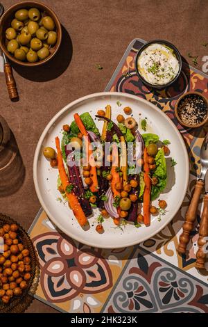 Salad with baked roasted carrots and chickpeas, lettuce, red onion, sesam, olive oil, green olives on ceramic plate on brown tablecloth, sunny light Stock Photo