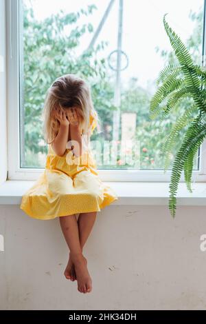 Sad lonely little girl sitting on windowsill crying and covering eyes with hands Stock Photo