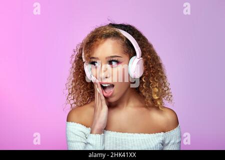 Shocked African American curly haired female listening to songs in headphones while expressing astonishment with opened mouth on gradient pink background in studio Stock Photo