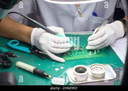 Close-up technician female on rubber gloves and work suit is using soldering to weld electronics circuit board on office desk Stock Photo