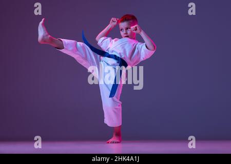 Portrait of sportive kid, male taekwondo, karate athletes in doboks doing basic movements isolated on purple background in neon. Concept of sport Stock Photo