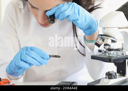 Woman scientist looking through magnifying glass at computer chip near microscope in laboratory Stock Photo