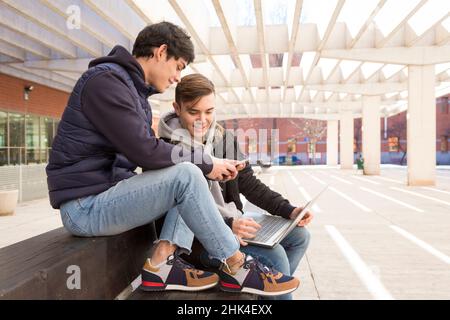 side view of two young university students sitting on a bench with laptop looking at a mobile phone