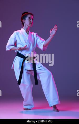 Portrait of female taekwondo, karate athletes in doboks doing basic movements isolated on purple background in neon. Concept of sport, martial arts Stock Photo