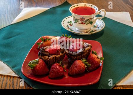 Double chocolate cheesecake cupcakes and strawberries with holly berry dinnerware, a cup of Christmas tea on a green napkin. Punch in the teacup. Stock Photo