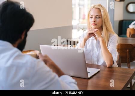 Close-up view from back of thoughtful attractive blonde woman sits at table with laptop in front of unrecognizable businss man drinking coffee. Stock Photo