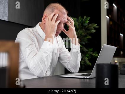 Tired or upset mature businessman sitting at desk and working on laptop. Employee thinking of deadline or workload or suffering from headache. Work overload concept. High quality photo Stock Photo