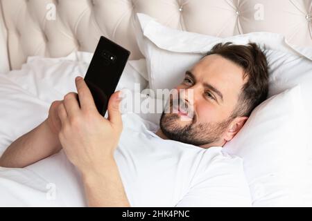 Handsome young man in bed typing on cell phone. Smiling caucasian man using mobile phone, browsing internet and texting on phone lying in bed at home. Stock Photo
