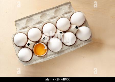 Ten white eggs in a cardboard tray, one with open shell showing the yellow egg yolk, light wooden background with copy space, high angle view from abo Stock Photo