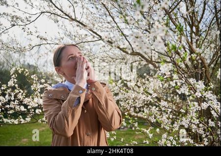 Woman allergic suffering from seasonal allergy at spring, posing in blossoming garden at springtime. Young woman sneezing in front of blooming tree. Spring allergy concept Stock Photo