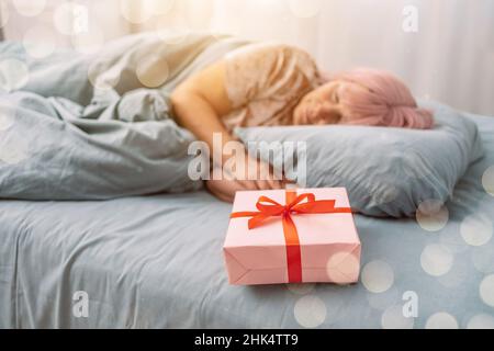 Woman in red sleep mask sleeping in bed near paper gift box with red satin ribbon decor. Christmas, New Year, Valentine's Day and birthday concept Stock Photo