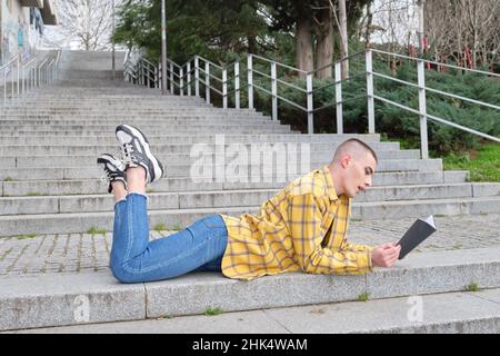 Young non binary person lying on stairs and reading a book. Stock Photo