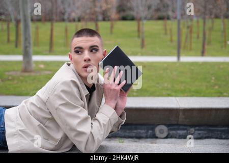 Young man wearing make up laying on a wall looking back with a book. Stock Photo