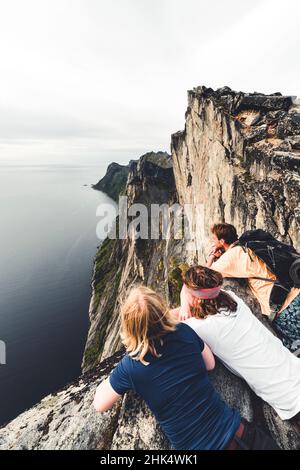 Three friends enjoying the view of the fjord leaning on rocks on Segla mountain peak, Senja island, Troms county, Norway, Scandinavia, Europe Stock Photo