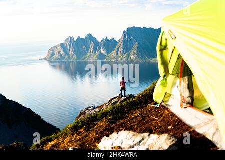 Hiker admiring mountains standing on top of cliff beyond camping tent, Senja island, Troms county, Norway, Scandinavia, Europe Stock Photo