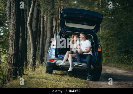 Young couple, man and woman, hugging together on a picnic, sitting in the trunk of a car in the woods, happy together Stock Photo