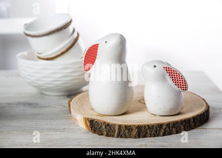 Two figurines of white hares with red ears big and small on a wooden saw cut and a bowl with eggs in the background in a white Scandinavian style kitc Stock Photo