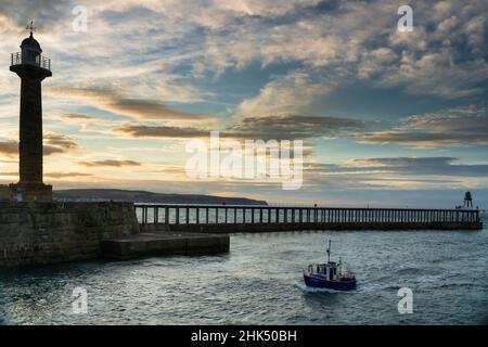 Sunset over Whitby harbour, pier and lighthouse as a small fishing boat comes into the harbour, Whitby, Yorkshire, England, United Kingdom, Europe Stock Photo